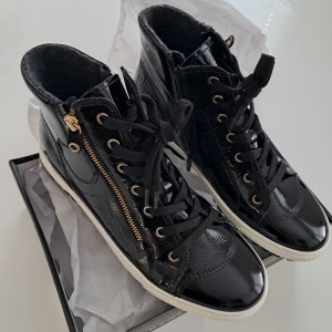 Svarta sneakers från Gabor med dragkedja - Snygga svarta sneakers från Gabor i glansigt skinn med svarta snören, guldfärgade detaljer och dragkedja på sidan. Insidan är fodrad med mjukt grått material för extra komfort. Yttersulan är vit med svart undersida och har bra grepp.