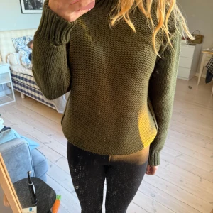 Olivgrön stickad tröja med knappar - Säljer en olivgrön stickad tröja med grov struktur och ribbade muddar. Tröjan har lång ärm, hög hals och snygga knappar på axeln och i sidan. 