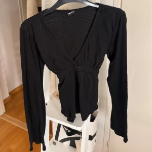 Svart omlottopp från Gina Tricot XS - Svart omlottopp från Gina Tricot i storlek XS. Toppen har lång ärm, v-ringning och knytband i ryggen för en snygg passform. Tröjan har även lite större ärmar! Den är i bra skick använd ett par gånger bara.