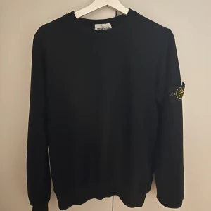 Svart sweatshirt från Stone Island - Svart sweatshirt från Stone Island med klassisk rund hals och ikonisk logotyp-patch på vänster ärm. Tröjan har lång ärm och ribbade muddar vid ärmslut och nederkant. Perfekt för en clean och stilren look.