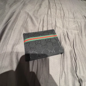 Svart plånbok från GG med ränder - Snygg svart plånbok från GG med diskret GG-mönster och klassiska gröna och röda ränder på framsidan. Insidan har flera fack för kort och sedlar, och är gjord i svart skinn och textil. Perfekt för dig som gillar stilrena accessoarer med lyxig känsla.