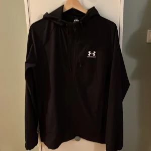 Svart vindjacka från Under Armour - Svart vindjacka från Under Armour med huva och dragkedja framtill. Jackan har en stilren look med logga på bröstet och meshfoder på insidan. Perfekt för dig som gillar sportig stil och vill ha något lätt och funktionellt.