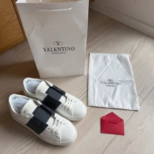 Valentino Garavani Open sneakers vit/svart - Snygga Valentino Garavani Open sneakers i vitt skinn med bred svart rem över ovansidan. Klassisk rund tå, platt sula med coola nitar längs kanten och snörning. Kommer med originalpåse och shoppingbag. Perfekta för dig som gillar exklusiv streetstyle.