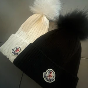 Moncler mössa med pälsboll - Två ribbstickade Moncler mössor, en vit och en svart, båda med stor fluffig pälsboll på toppen och Moncler-logga framtill. Dock den svarta mössan så är bollen äkta päls men inte den vita! Perfekt för dig som vill ha en snygg och lyxig vinteraccessoar med streetkänsla. 300kr för den vita 500 för den svarta! 