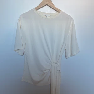 Beige knytblus från H&M - Säljer en stilren gulvit blus från H&M med korta ärmar och rund hals. Blusen har en snygg knytning som man även kan välja hur man knyter . Materialet känns mjukt och lätt. Aldrig använt!💕