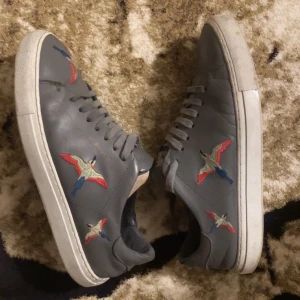 Grå Axel Arigato - Snygga grå sneakers från Axel Arigato med broderade fåglar i rött, blått och beige på sidorna. Skorna har snörning och en vit platt sula. Tillverkade i skinn för en lyxig känsla och stilren look. Perfekt för dig som gillar detaljer och vill sticka ut.