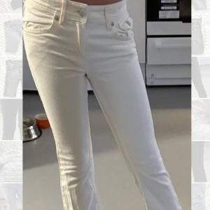 Vita jeansbyxor med fickdetaljer - Snygga vita jeansbyxor med raka ben och coola fickdetaljer både fram och bak. Byxorna har hög midja och klassisk femficksdesign med extra lock på fickorna. Perfekta för dig som vill ha en clean och fräsch look.