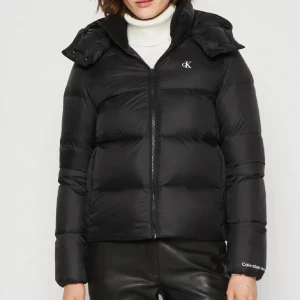 Svart pufferjacka från Calvin Klein - Säljer en svart pufferjacka från Calvin Klein med huva och dragkedja framtill. Jackan har en stilren design med CK-logga på bröstet och Calvin Klein Jeans-brodyr vid ärmslutet. Perfekt för kalla dagar och har en skön, vadderad känsla.