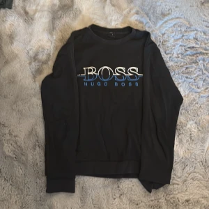 Svart Hugo Boss tröja med logga - Svart långärmad tröja från Hugo Boss med stor vit och blå logga på bröstet. Slim fit-modell med ribbade muddar vid ärmslut och nederkant. Perfekt för dig som gillar stilrena och klassiska plagg med en modern touch.