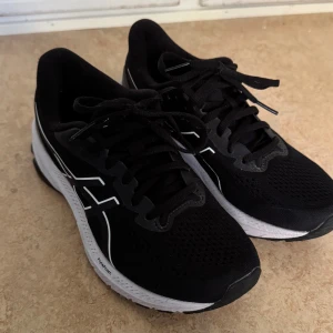 Svarta Asics sneakers med vit sula - Säljer ett par svarta Asics sneakers med vit och svart sula. Skorna har snörning, ventilerande mesh-ovandel och klassisk Asics-logga i vitt på sidorna. Perfekta för dig som gillar sportig stil och vill ha bekväma skor till vardagen. Använd max 3 gånger. Storlek 37,5