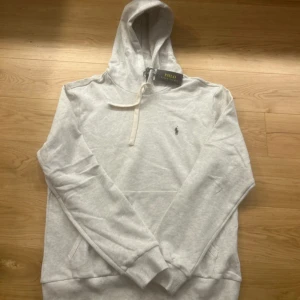 Ljusgrå hoodie från Polo Ralph Lauren - Säljer en stilren ljusgrå hoodie från Polo Ralph Lauren med klassisk broderad logga på bröstet. Tröjan har huva med snörning, half zip-dragkedja och känguruficka. Perfekt för dig som gillar en clean och avslappnad look. Hoodien kan bäras av en person som är M men liten större.