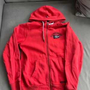 Röd hoodie från Peak Performance - Röd hoodie från Peak Performance med dragkedja, huva med snörning och broderad logga på bröstet. Mjuk bomullskänsla och två stora fickor framtill. Perfekt för en avslappnad och sportig stil.