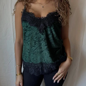 Grönt sammetslinne med spetsdetaljer - Mörkgrönt spetslinne i mjuk sammet med svarta spetsdetaljer längs urringning och nederkant. Tunt axelbandslinne med avslappnad passform, perfekt för att lyfta vilken outfit som helst.