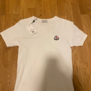 Vit märkes t-shirt - Snygg vit t-shirt med klassisk rund hals och korta ärmar. På bröstet sitter Monclers broderade logga i svart, rött och blått. Materialet är mjuk bomull som känns skönt mot huden. Perfekt för dig som gillar stilrena designer och exklusiva märken.