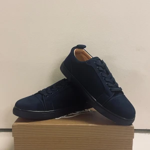 Mörkblå sneakers från Christian Louboutin - Snygga mörkblå sneakers från Christian Louboutin med klassisk röd sula och diskret logga på hälen. Skorna har snörning, rund tå och är tillverkade i mocka med detaljer i skinn. Perfekta för dig som vill ha en clean och lyxig look.