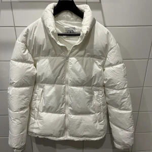 Vit pufferjacka från Zara, storlek M - Snygg vit pufferjacka från Zara i storlek M. Jackan har hög krage, dragkedja framtill och två fickor på sidorna. Perfekt för kyliga dagar och ger en clean look. Tillverkad i syntetmaterial och har en klassisk pufferdesign.