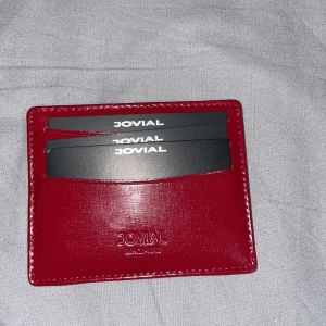 Röd plånbok från Jovial i skinn - Slimmad röd plånbok från Jovial i äkta skinn, perfekt för kort och sedlar. Snyggt präglad logga framtill och flera fack för enkel organisering. Kommer i en lyxig brun presentask med gulddetaljer. Stilren och smidig att ha med sig överallt.