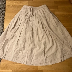 Beige långkjol med veckad design - Kjolen är 70 cm lång.                                            Använd bara en gång, i nyskick.                         Luftig och snygg beige långkjol med veckad design och resår i midjan. Kjolen har en lätt A-linjeform och är tillverkad i ett tunt bomullstyg som ger en skön känsla. Perfekt för dig som gillar en stilren och enkel look.