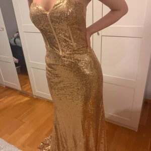 Guldig glittrig långklänning - Säljer en superglammig guldig långklänning med paljetter över hela tyget. Klänningen är axelbandslös, figurnära och har en snygg släp. Perfekt för dig som vill sticka ut och glänsa på bal eller fest. Dragkedja i ryggen för enkel påklädning.