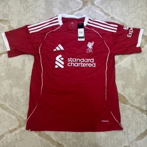 Liverpool röd fotbollströja Adidas L - Liverpool FC matchtröja från Adidas i rött med vita detaljer och tryck. Klassisk design med klubbmärke, sponsorlogga och tre vita ränder på axlarna. Tillverkad i lätt och ventilerande polyester med Aeroready-teknologi. Slim fit-modell med korta ärmar.