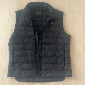 Svart dunväst från Morris, medium - Mörkblå Norfolk liner vest från Morris i storlek medium. Västen har ståkrage, dragkedja framtill och två fickor med dragkedja. Klassisk och stilren design med diskret Morris-logga på bröstet. Perfekt lager-på-lager-plagg för kyligare dagar.