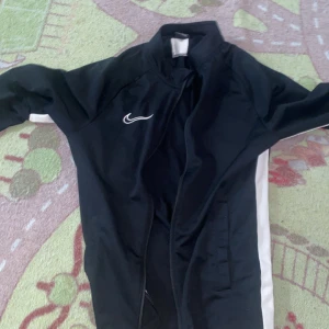 Nike svart träningsjacka Dri-Fit S - Svart träningsjacka från Nike i Dri-Fit-material med vita detaljer längs sidorna och ärmarna. Jackan har dragkedja framtill, ståkrage och två fickor. Perfekt för dig som gillar sportig stil och vill ha något bekvämt till träningen.