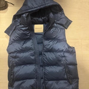 Mörkblå dunväst från GANT med huva - Snygg mörkblå dunväst från GANT med avtagbar huva och två praktiska fickor med dragkedja. Västen har quiltad design och ett diskret GANT-märke på bröstet. Perfekt för lager-på-lager och chill stil under höst och vinter. Använd 1 gång sen köpt i butik. Nypris 2699. Säljer då den bara tar plats i garderoben.
