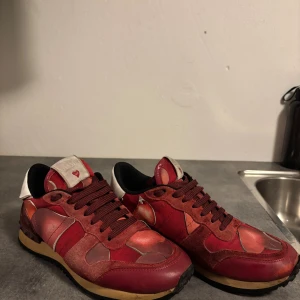 Valentino Rockrunner röda sneakers hjärtan - Unika Valentino Rockrunner sneakers i rött med hjärtmönster. Skorna har detaljer i mocka, skinn och textil, samt klassisk snörning och svart yttersula. Vit detalj på hälen och ikoniska nitar baktill. Perfekta för dig som vill sticka ut med cool design.