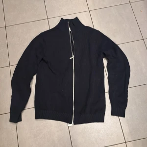 Mörkblå stickad kofta med dragkedja - Säljer en mörkblå stickad kofta med hel dragkedja framtill och hög krage. Tröjan har lång ärm och ribbade muddar vid ärmslut och nederkant. Perfekt för lager-på-lager och chill stil.