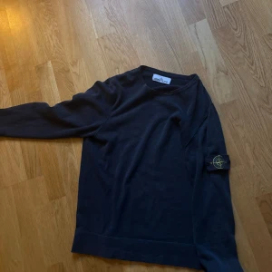 Mörkblå Stone Island stickad tröja - Mörkblå stickad tröja från Stone Island med klassisk patch på vänster ärm. Tröjan har rund halsringning och långa ärmar. Perfekt för dig som gillar streetwear och vill ha något stilrent med ikonisk logga.