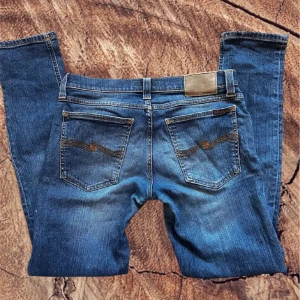 Nudie Jeans Gim Tim - Säljer ett var blåa grim tim jeans från Nudie Jeans med snygg tvätt, slim passform och raka ben. Jeansen har fem fickor, knappgylf och snygga slitningar framtill. W30 L32. Är lagade i skrevet men det syns inte när man har dem på.