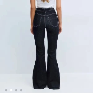 Mörka bootcut jeans med kontrastsömmar - Snygga mörkblå jeans med bootcut passform och hög midja. Jeansen har tydliga vita kontrastsömmar, klassiska fem fickor och stängs med knapp och dragkedja. Perfekta för dig som gillar en retroinspirerad look med extra vidd nertill.