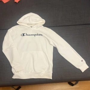 Vit hoodie från Champion med logga - Säljer en vit hoodie från Champion med stor svart logga på bröstet och liten logga på ärmen. Tröjan har huva, magficka och långa ärmar. Materialet är mjukt och består av bomull och polyester, perfekt för chill dagar. Klassisk och clean stil.