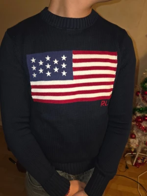 Mörkblå stickad tröja Polo Ralph Lauren - Säljer en mörkblå stickad tröja från Polo Ralph Lauren med amerikansk flagga på bröstet och RL-broderi i rött. Tröjan har rund halsringning, ribbade muddar och är långärmad. Perfekt för dig som vill ha en klassisk och snygg statement piece.