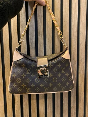 Brun  axelväska - axelväska i brunt med beigea detaljer och guldfärgad kedja. Väskan har ett snyggt lås framtill och insida med dragkedjefack. 