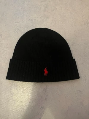 Svart mössa från Polo Ralph Lauren - Klassisk svart mössa från Polo Ralph Lauren i mjuk merinoull. Mössan har uppvikt kant med ribbad struktur och en röd broderad logga framtill. Perfekt accessoar för kyliga dagar och enkel att matcha med olika outfits.                        MÖTAS FÖR KÖPAS (Stockholm)