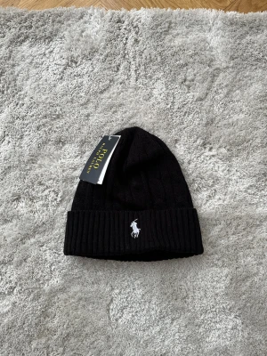 Svart kabelstickad mössa Polo Ralph Lauren - Kabelstickad svart mössa från Polo Ralph Lauren med klassisk vit broderad logga framtill. Mössan har bred ribbad kant och är tillverkad i mjukt stickat material som håller dig varm och snygg under kyliga dagar.