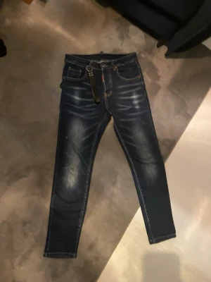 Mörkblå raka jeans från Dsquared2 - Säljer ett par helt nya mörkblå raka jeans från Dsquared2 med snygga slitningar och tvättade detaljer. Klassisk femficksmodell med Dsquared2-lapp bak i midjan och coolt läderband i hällan. Tillverkade i Italien i mjuk bomullsdenim.
