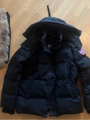 Svart dunjacka från Canada Goose - Säljer en svart dunjacka från Canada Goose med huva och klassisk logga på ärmen. Jackan är tjockt vadderad och har dragkedja framtill samt flera praktiska fickor. Perfekt för kalla vinterdagar och riktigt snygg streetstil.