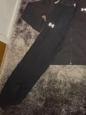 Svart träningsväst från Under Armour - Säljer en svart träningsväst från Under Armour med dragkedja framtill och logga på bröstet. Västen är i ett sportigt material som passar perfekt till träning och har en stilren, enkel design.