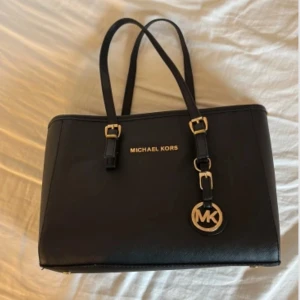 Svart handväska från Michael Kors - Snygg svart handväska från Michael Kors i skinn med guldfärgade detaljer och dubbla axelband. Väskan har dragkedja upptill, flera innerfack och ett klassiskt MK-hänge framtill. Perfekt storlek för allt du behöver ha med dig och stilren design.