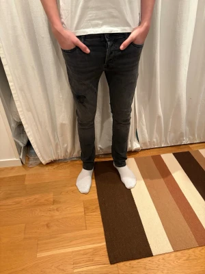 Svarta slim jeans med slitning - Säljer ett par svarta slim jeans med snygg slitning på ena låret. Jeansen har klassisk femficksdesign och smal passform hela vägen ner. Materialet är stretchigt för skön känsla och de har en mörk tvätt som passar till det mesta. Märket är Jack&Jones och storleken är 28/30