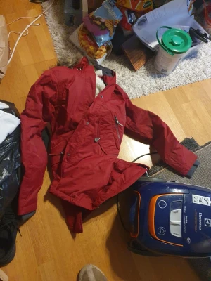 Röd parkas med fuskpäls - Säljer en röd parkas med huva och fuskpälsdetalj. Jackan har dragkedja och knappar framtill, flera fickor och är långärmad. Perfekt för kalla dagar och har en snygg, klassisk look.