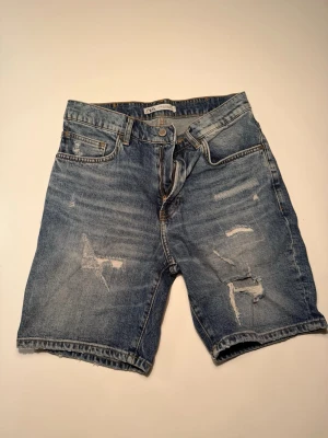Blå denimshorts från Zara - Säljer ett par blå denimshorts från Zara med slitna detaljer och råa kanter. Klassisk femficksmodell med knappgylf och bälteshällor. Perfekta för sommaren och har en avslappnad loose fit.