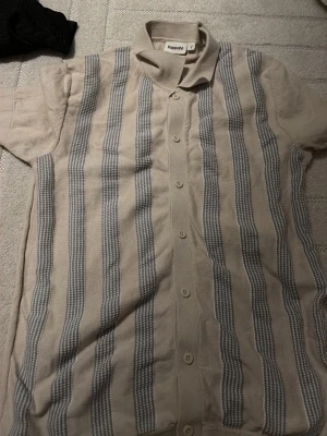 Randig kortärmad skjorta Kappahl - Kortärmad skjorta från Kappahl i beige med vertikala grå ränder. Skjortan har klassisk krage och knappar framtill. Materialet känns mjukt och luftigt, perfekt för varmare dagar. Snygg retrovibe och passar till många olika stilar.