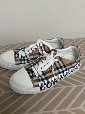 Burberry sneakers  - Snygga sneakers från Burberry med klassiskt rutigt mönster i beige, svart, vitt och rött. Skorna har vit sula, vita snören och stor Burberry-logga på sidan. Ovandelen är i textil och insidan i vitt skinn. Perfekta för dig som vill sticka ut med en ikonisk look. Lite sönder på sidan såsom man ser på bilden men inte riktigt något som sticker ut mycket. Storlek 42 
