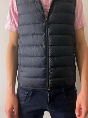 Svart dunväst UNIQLO - Säljer en svart dunväst med horisontella sömmar och hel dragkedja framtill. Västen har en enkel, stilren design och är lätt vadderad för extra värme. Perfekt att slänga över en hoodie eller t-shirt för en casual look.
