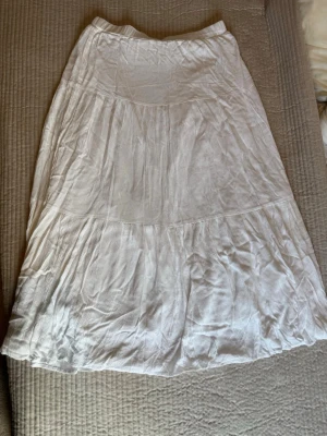 Vit långkjol i bomull - Säljer en vit långkjol i mjukt och luftigt bomullstyg. Kjolen har en volangig design med flera sömmar som ger ett flowy intryck. Perfekt för dig som gillar en enkel men ändå snygg look. 