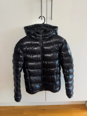 Svart pufferjacka från Colmar - Säljer en svart, glansig pufferjacka från Colmar med huva och dragkedja framtill. Jackan har Colmar-logga på ärmen och är vadderad för extra värme. Perfekt för kalla dagar och har en riktigt snygg streetstil.