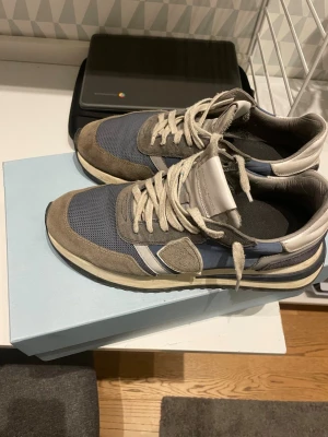 Philippe Model blå/grå sneakers - Säljer ett par Philippe Model sneakers i blått och grått med beige detaljer. Skorna har snörning, rund tå och platt sula. Ovandelen är i mocka och textil, med vita och grå inslag samt en snygg kontrast på sulan. Perfekta för dig som gillar stilrena och sportiga sneakers.